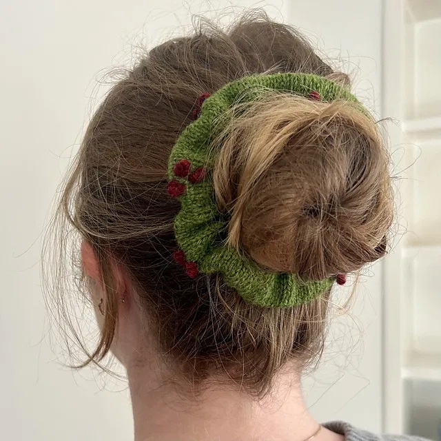 Nuca di ragazza con chignon avvolto da uno scrunchie verde ai ferri e decorato da bacche rosse a maglia. Il modello è di Hilly Knits.